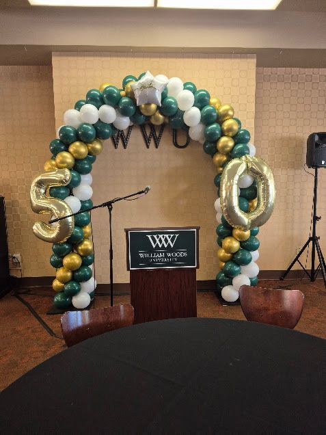Balloon arch in teal, gold, and white around a podium with microphones at an event stage