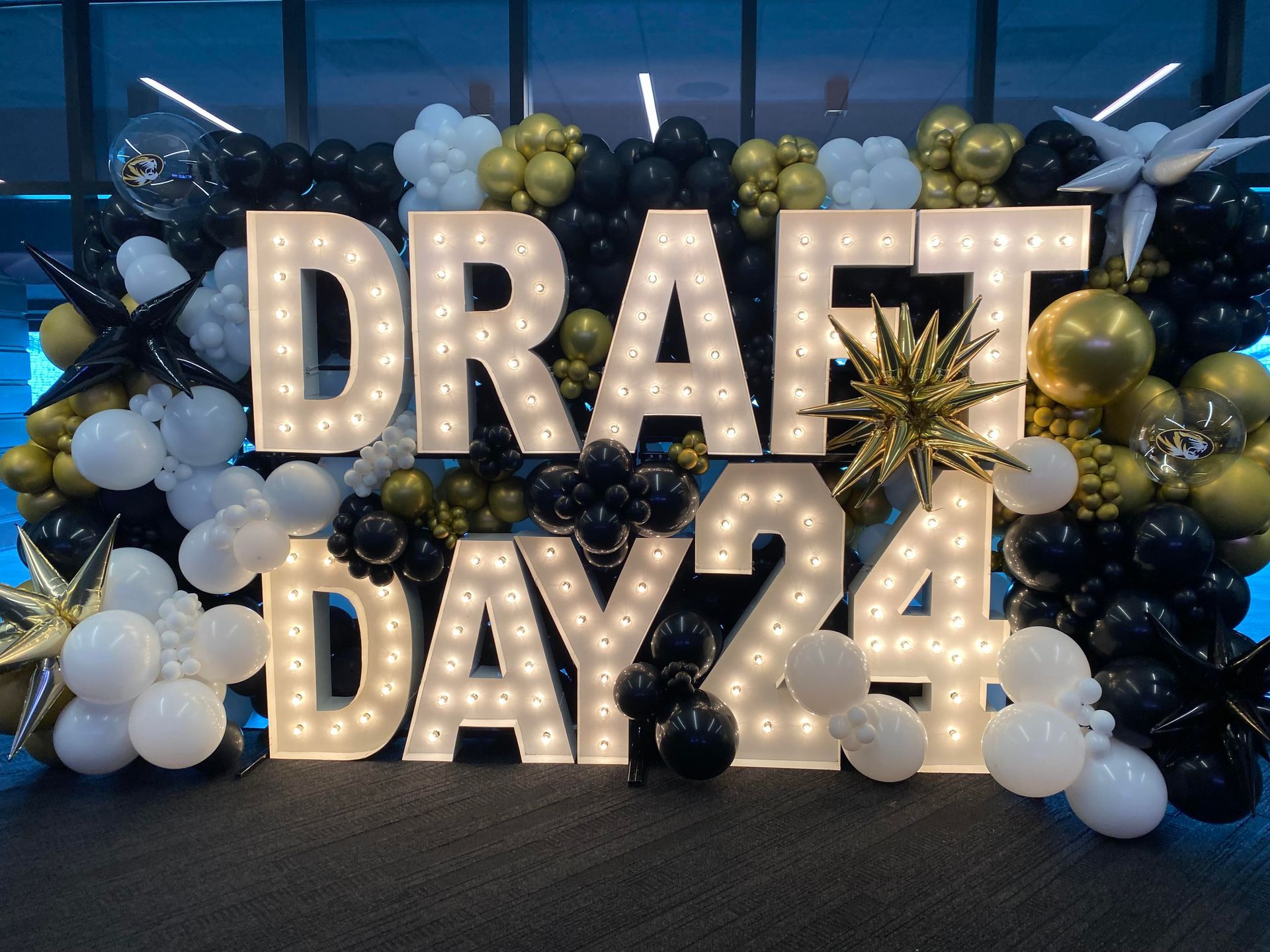 Marquee letters reading “DRAFT DAY 24” surrounded by blue, gold, and white balloons at an event backdrop