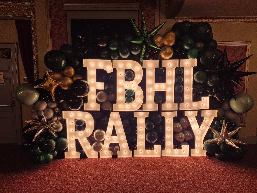 Lit marquee letters reading “FBHL RALLY” against a backdrop of balloons and greenery