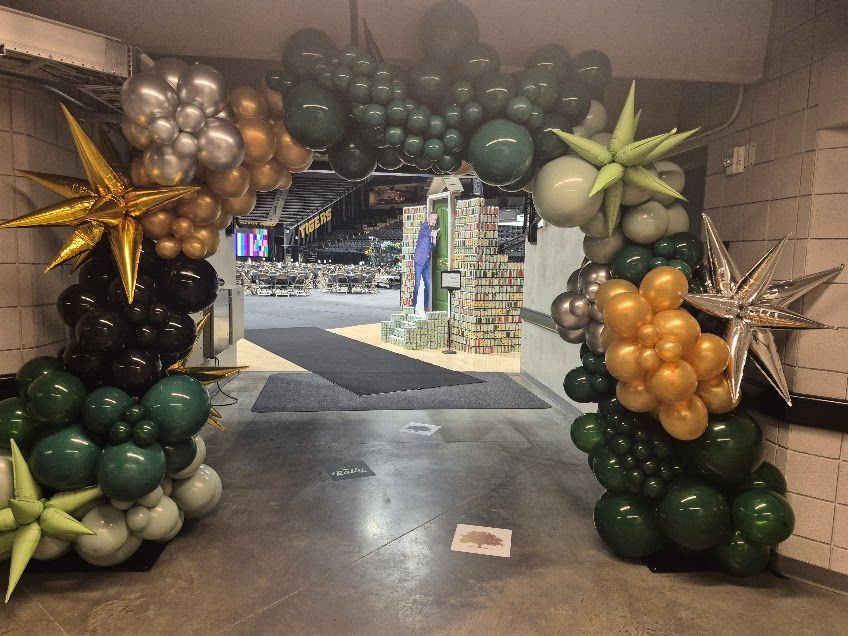 Balloon arch in gold, silver, green, and black framing a walkway entrance outdoors