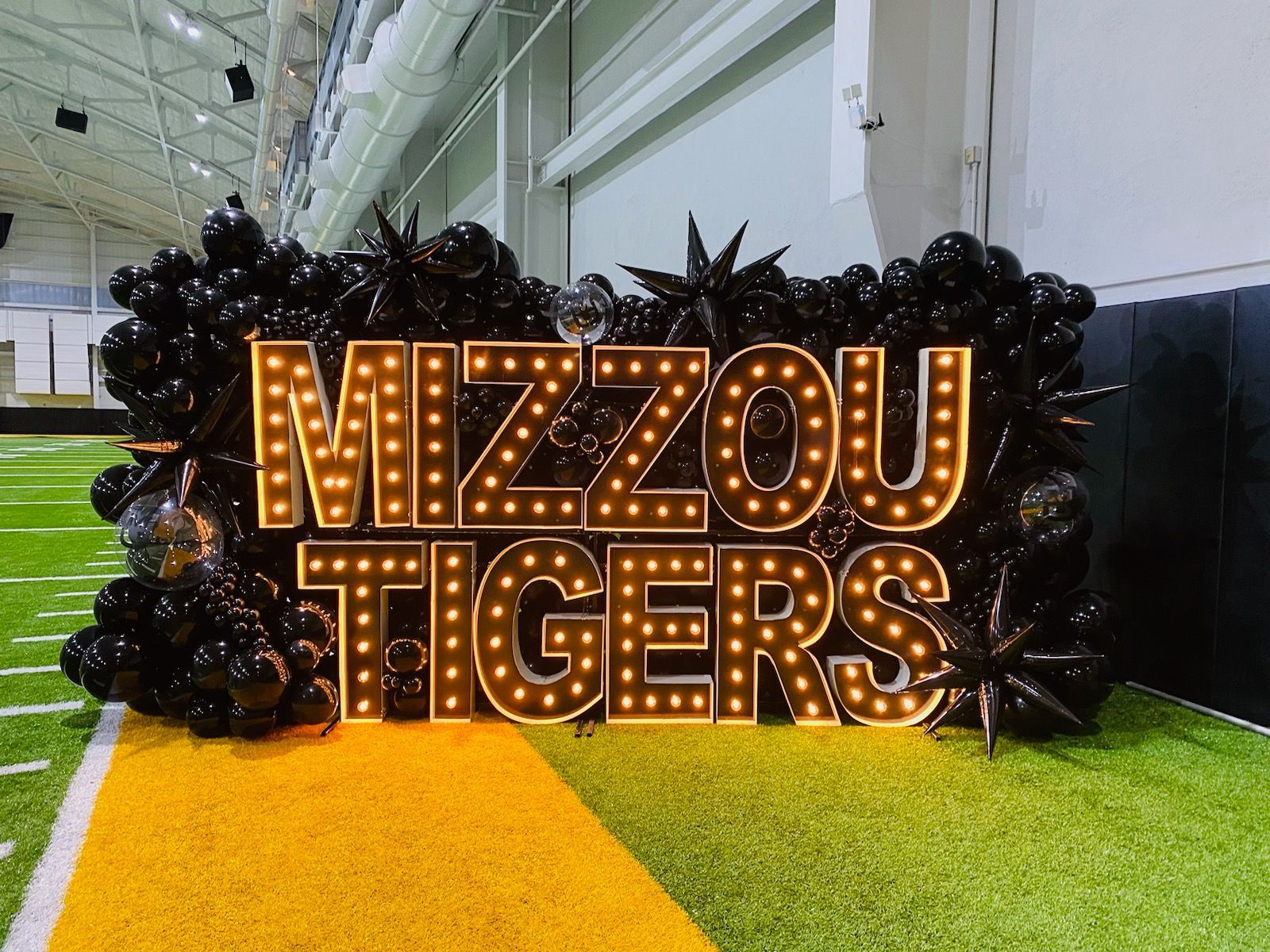 Lit sign reading “MIZZOU TIGERS” with black tiger decor on a yellow turf display indoors