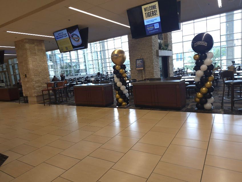 Airport lobby with tiled floor, check-in counters, and black-and-gold balloon columns near windows