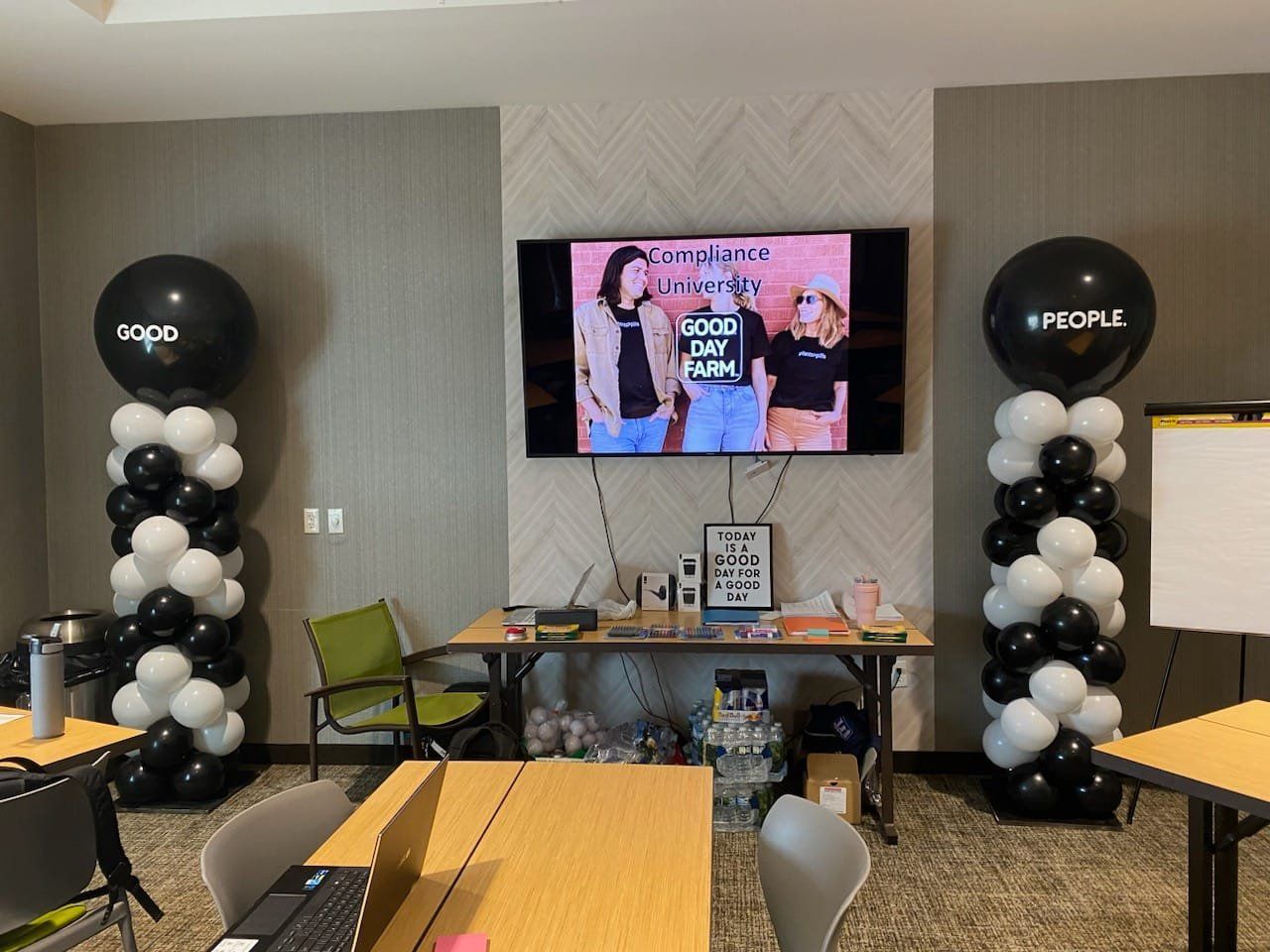 Decorated event space with balloon columns, tables, and a wall-mounted TV displaying a celebration slide.
