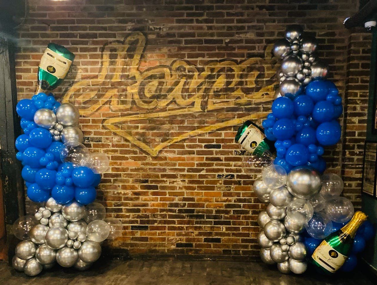 Blue and silver balloon display with champagne bottles against a brick wall backdrop