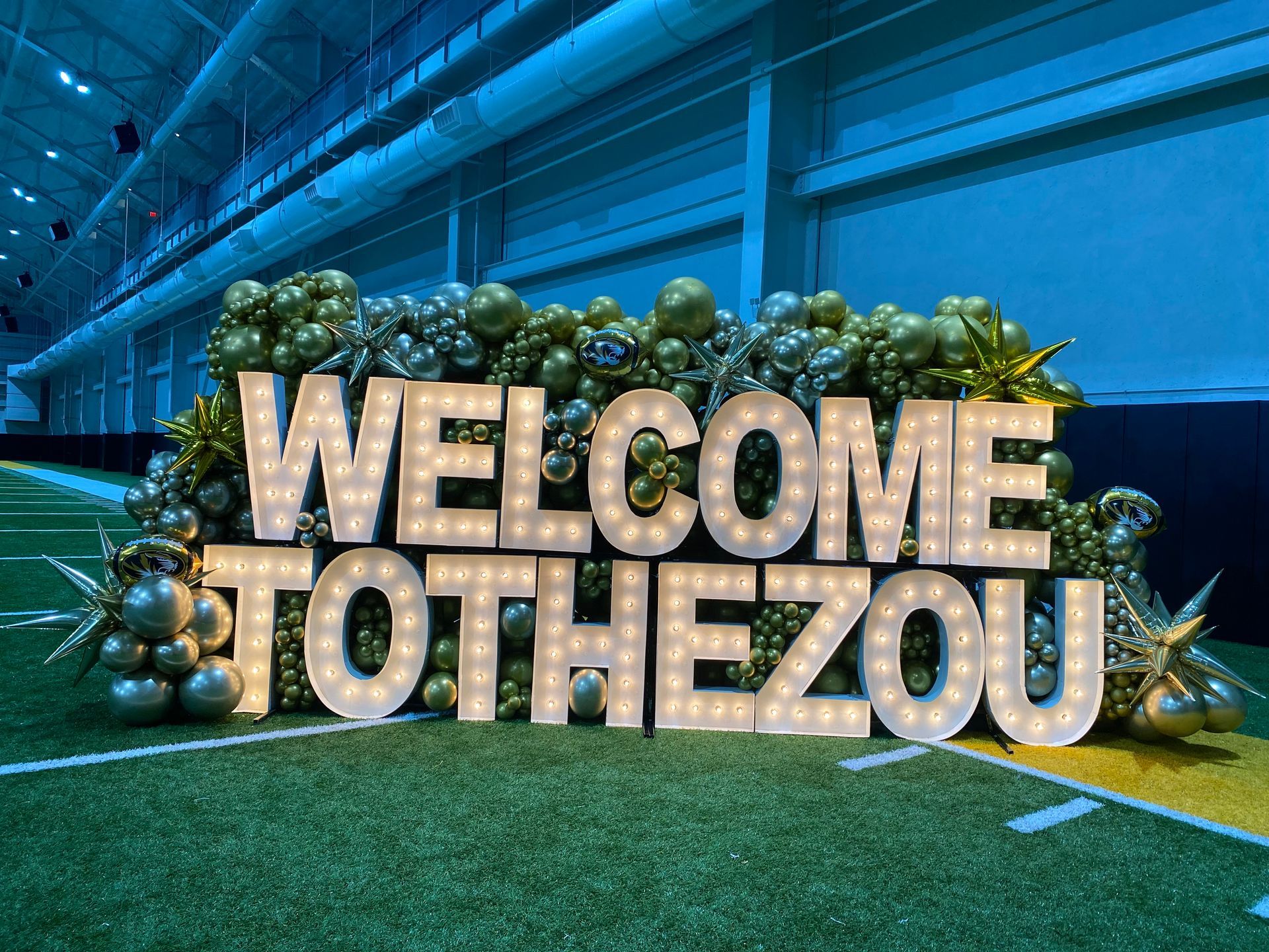 Welcome to the Zoo sign with gold letters, greenery, and ornaments in a blue-lit indoor display.