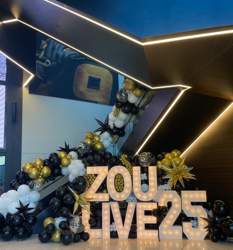 Gold “ZOU LIVE25” sign with black, white, and gold balloon decor in a modern indoor setting