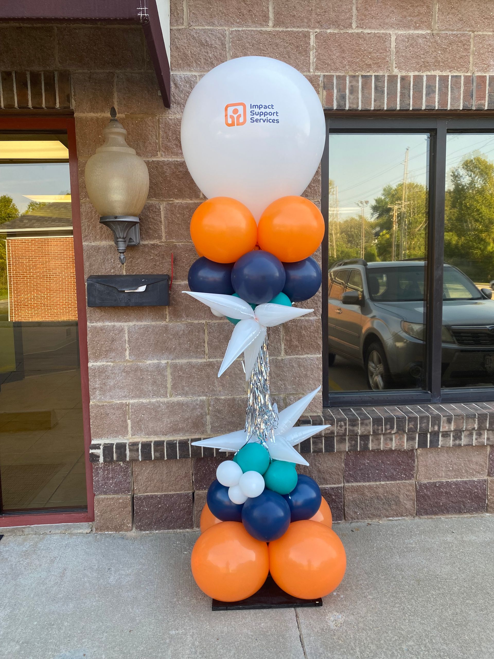 Balloon column with white, orange, and navy balloons beside a brick wall and window entrance
