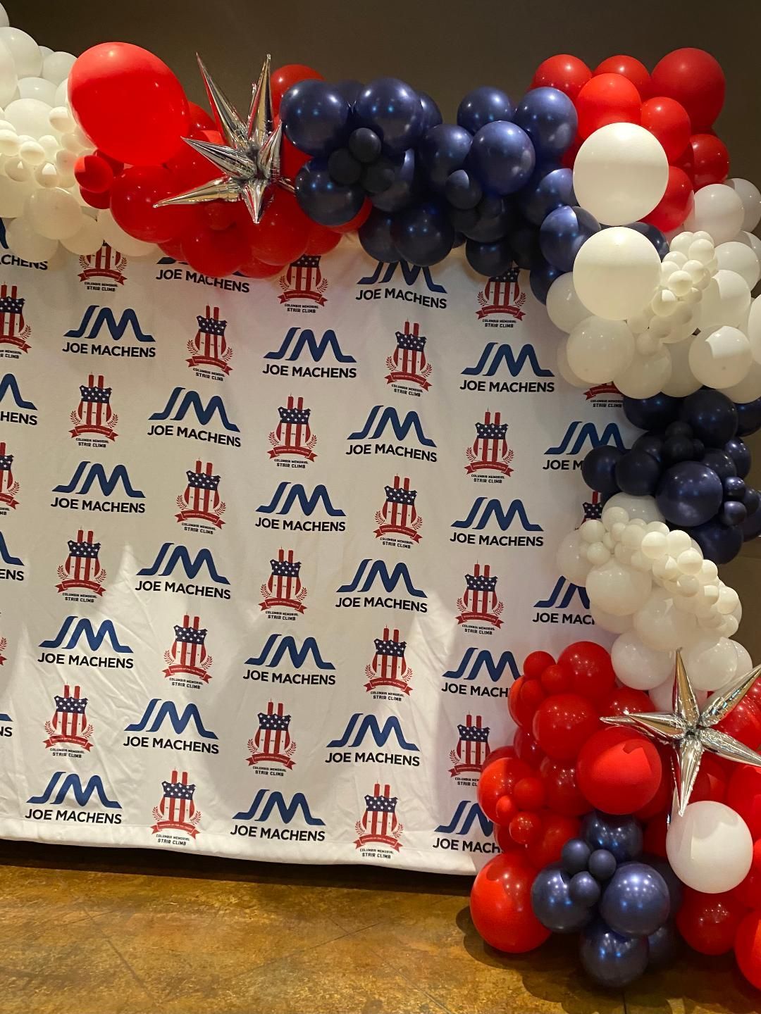 Red, white, and blue balloon arch beside a branded step-and-repeat backdrop with JM logos