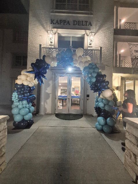 Balloon arch framing a lit café entrance at night, with blue, white, and gold balloons