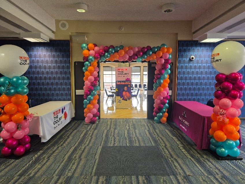 Colorful balloon arch framing a doorway in a decorated indoor hallway with tables on both sides