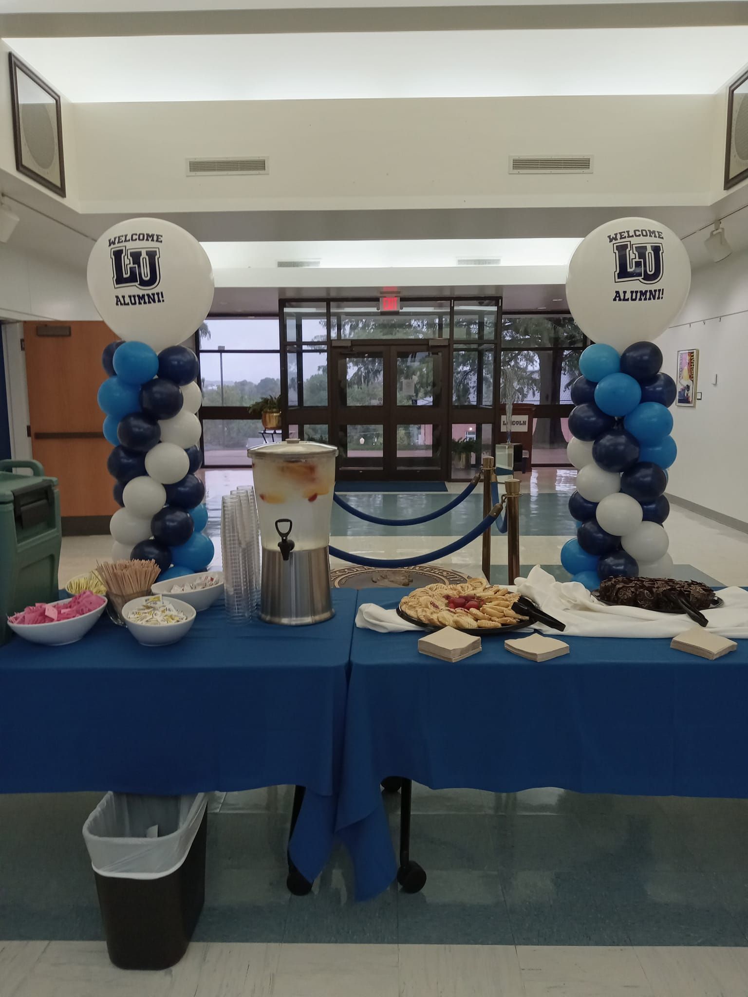 Decorated blue table with cakes and treats at an indoor event, flanked by white and blue balloon columns