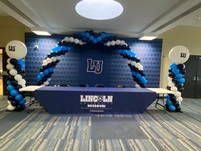 Lincoln University event backdrop with blue-and-white balloon arch and podium table in a conference room