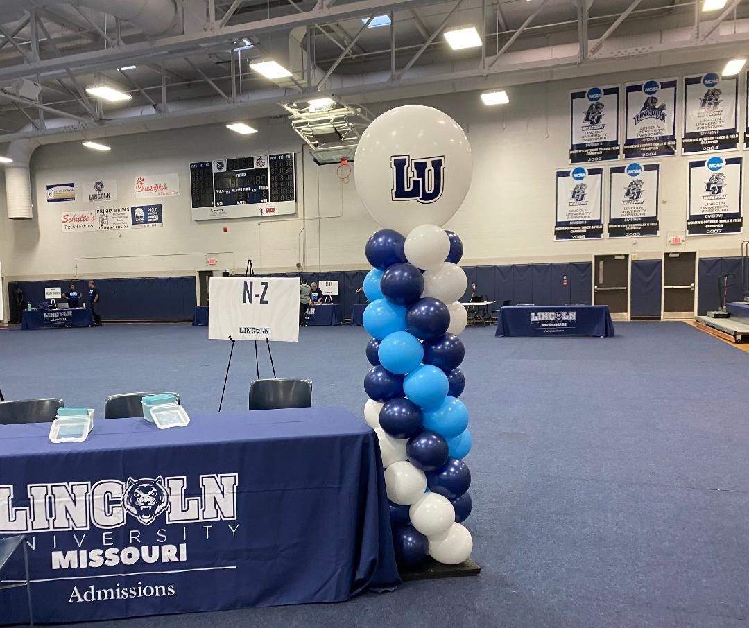Mizzou recruiting setup in gym with blue-and-white balloon column and table banner