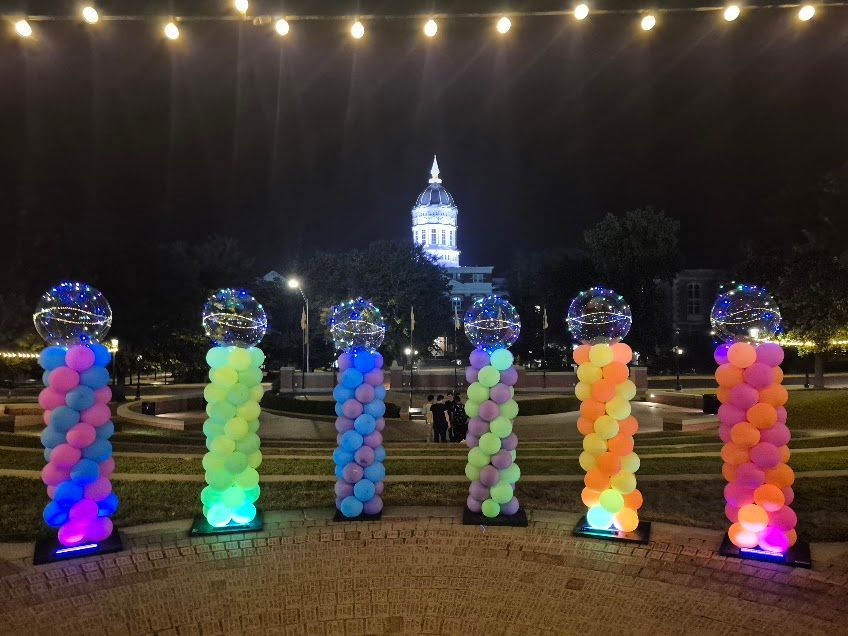 Colorful balloon columns lit at night with a glowing building in the distance