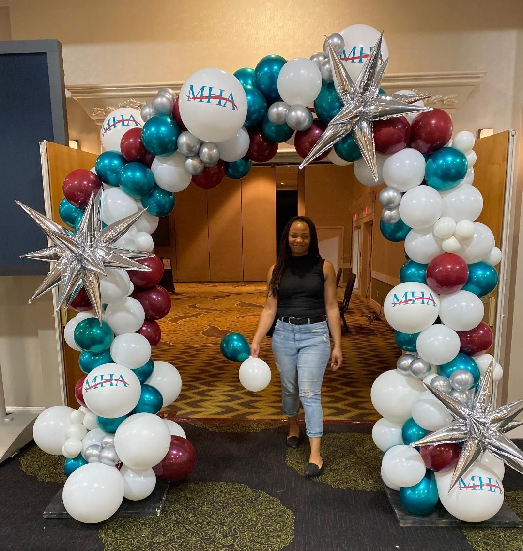 Person standing under a teal, white, and burgundy balloon arch at an indoor event