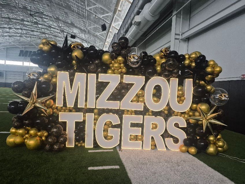 “MIZZOU TIGERS” gold sign with black and gold balloons in an indoor turf area