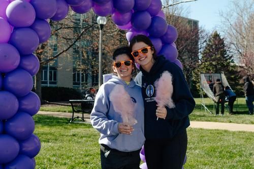 Two people posing under a purple balloon arch on a sunny lawn, smiling and wearing sunglasses