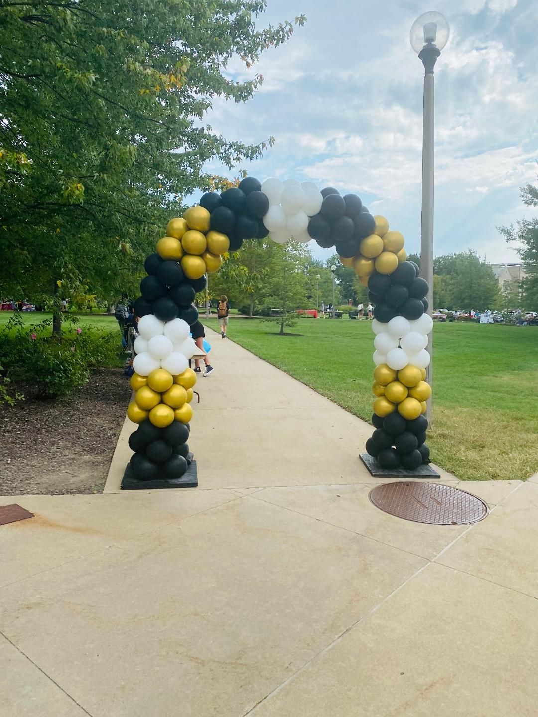Black, gold, and white balloon arch on a park walkway with trees and lamp post nearby