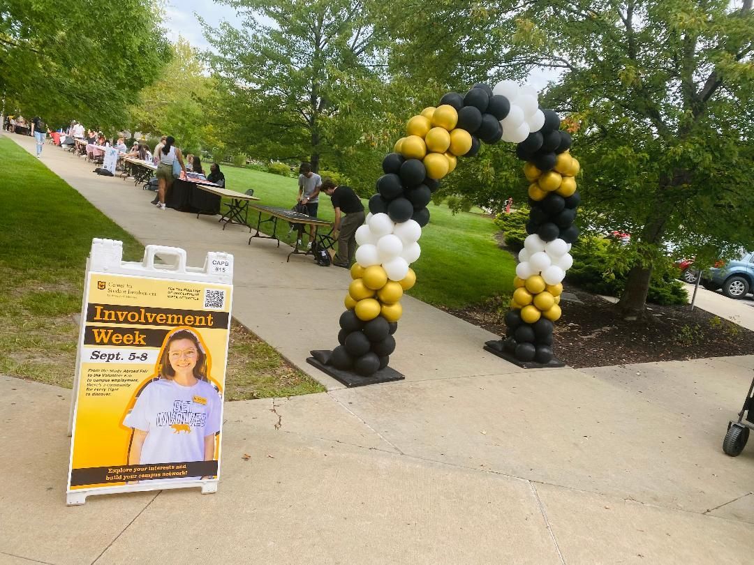 Yellow and black balloon arch at an outdoor event with tables and a welcome sign