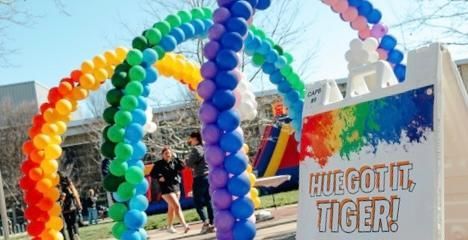 Rainbow balloon arches at an outdoor event beside a sign that reads “HUEGOTTE TIGER!”