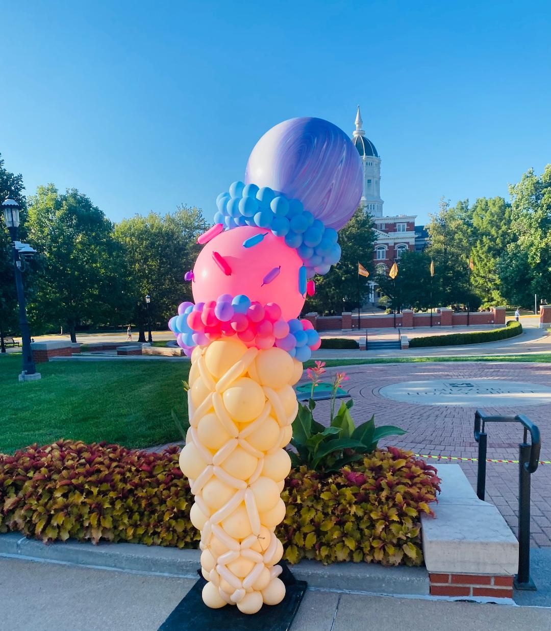 Tall colorful balloon sculpture shaped like an ice cream cone in a park with trees and a building in the background