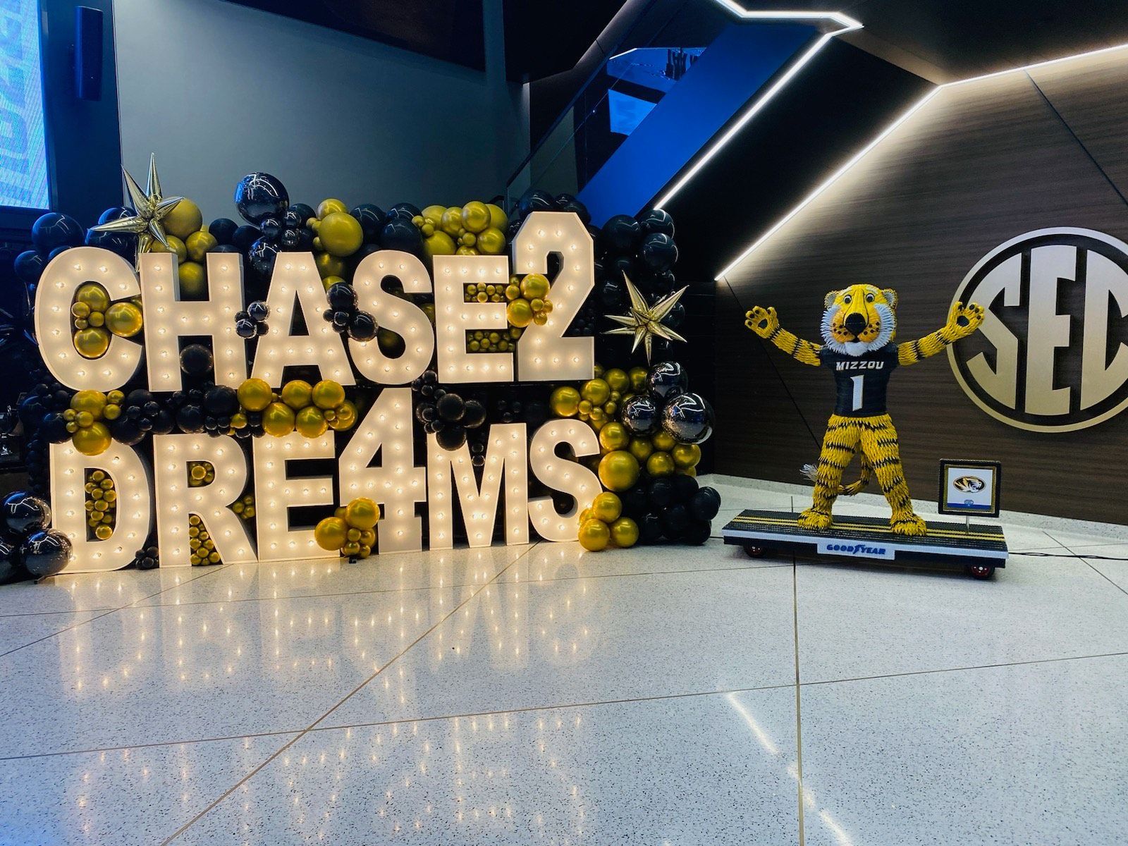 College event display with gold “CHASE 2 DRE4MS” letters, blue and gold balloons, and a mascot figure holding pom-poms.
