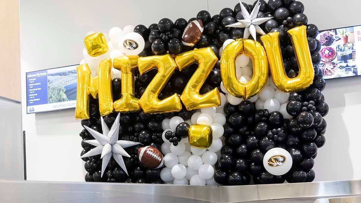 Gold “Mizzou” balloon sign on a black, white, and silver balloon display in an indoor lobby