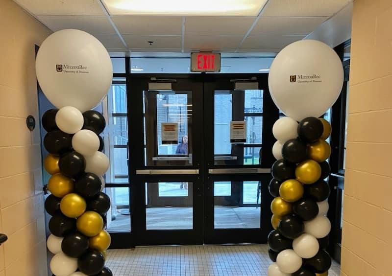 Balloon columns in black, white, and gold framing a building entrance with glass doors.