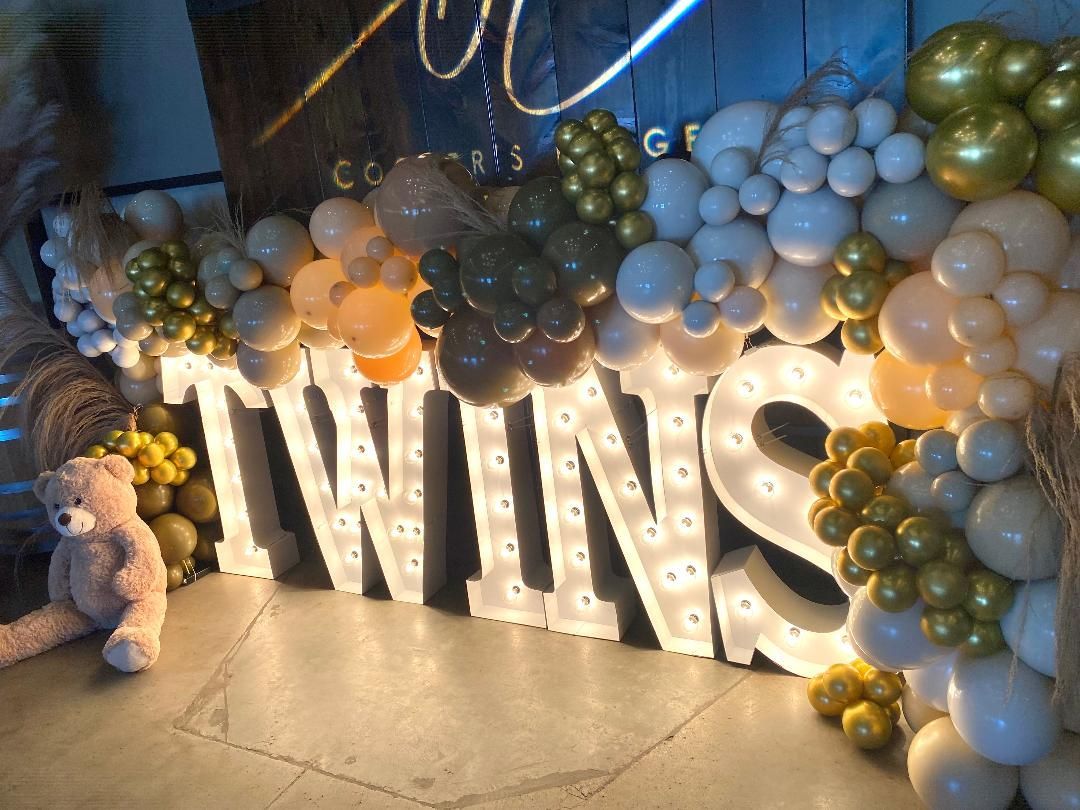 Lit “TWINS” sign surrounded by white, green, and gold balloon garland at a celebration backdrop