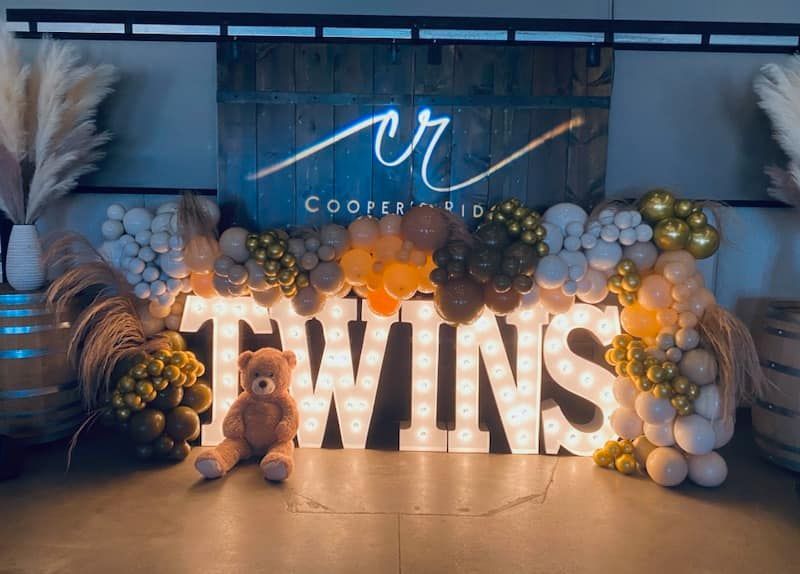 Decorative twin announcement display with large lit “TWINS” letters and balloon arch in blue, gold, and white