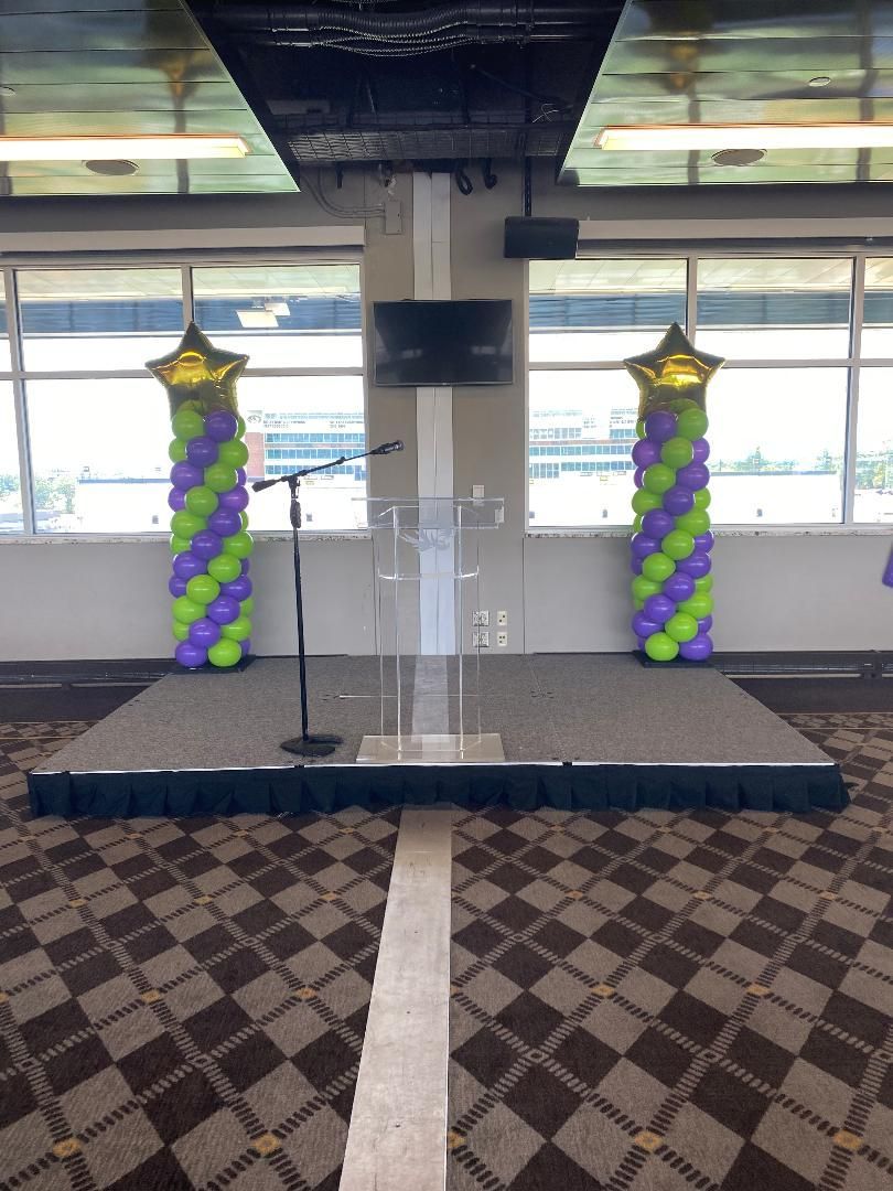 Decorated indoor stage with black platform, clear podium, and purple-green balloon columns by windows