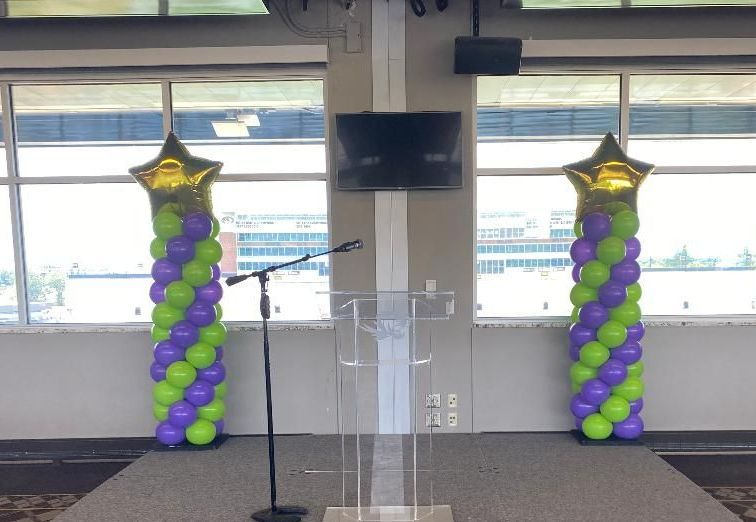 Clear podium on stage with black microphone stands and purple-green balloon columns by large windows.