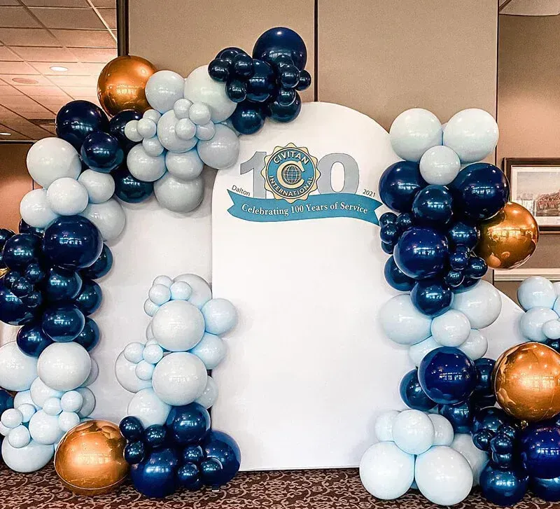 White event backdrop framed by navy, light blue, and gold balloon garlands with a “100” sign at the top