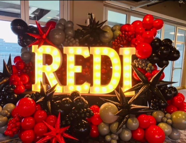 Illuminated REDI sign surrounded by red, black, and gold balloons in an indoor display