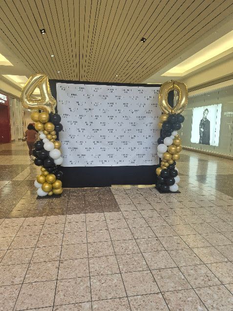 Black, gold, and white balloon arch framing a step-and-repeat backdrop in a shopping mall.
