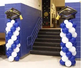 Blue and white balloon columns flank a stairway entrance with graduation caps on top.