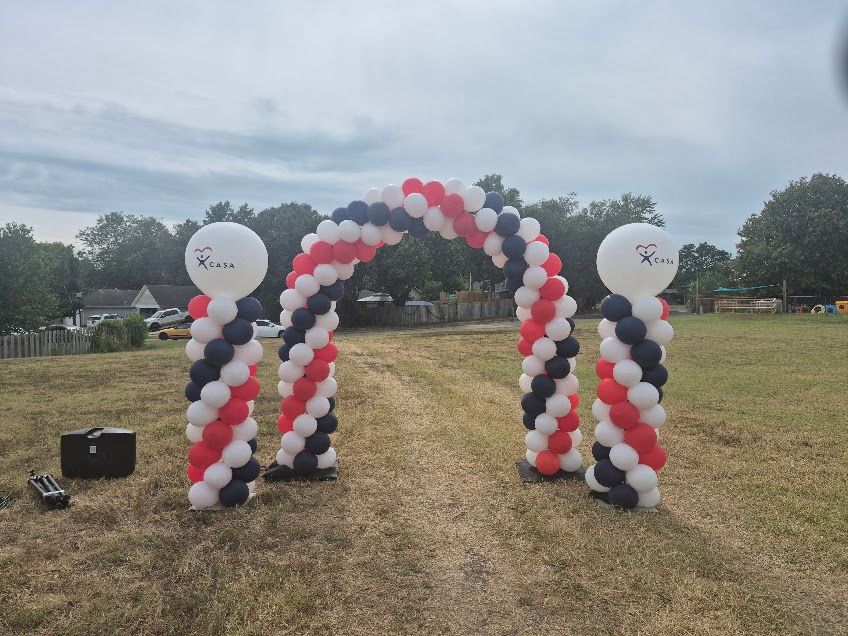 Red, white, and black balloon arch on a grassy field with two white balloon columns and balloons.