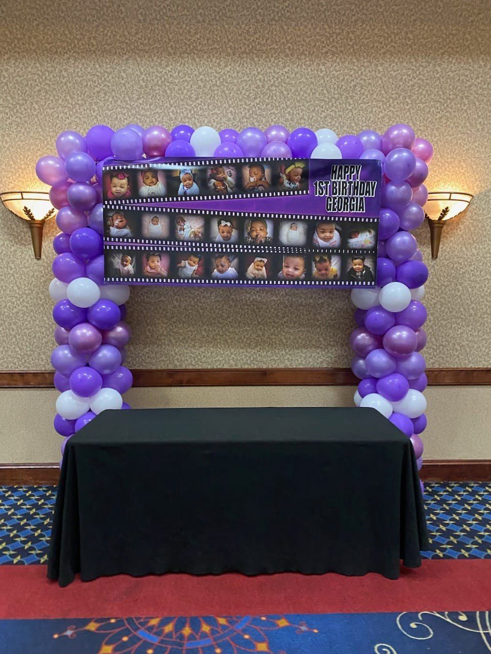 Purple balloon arch framing a black table in a hallway with a poster display above it