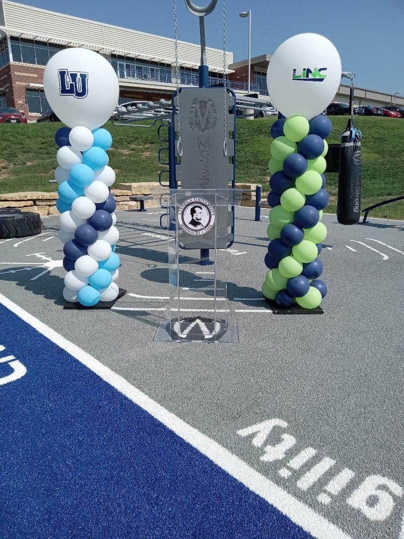 Two balloon columns flank a clear lectern on a blue track at a stadium entrance.