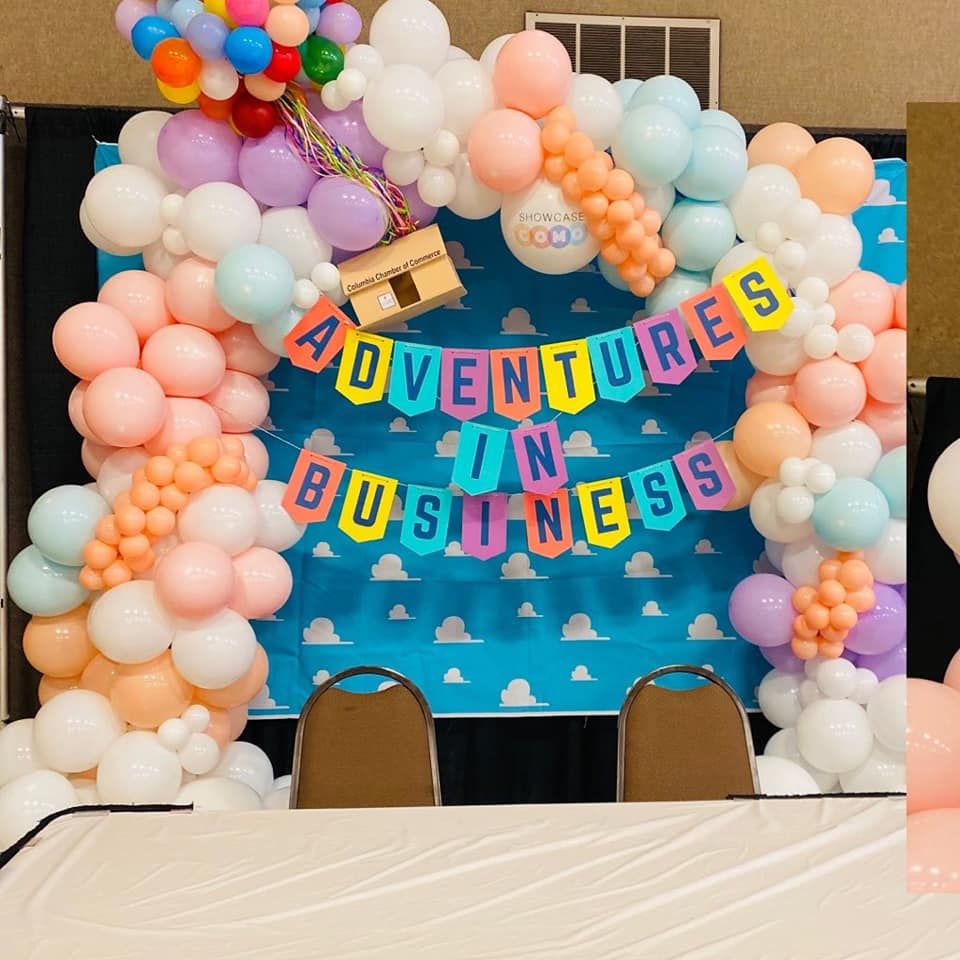 Colorful balloon arch and “Adventures in Business” banner on a blue backdrop with chairs and table in front