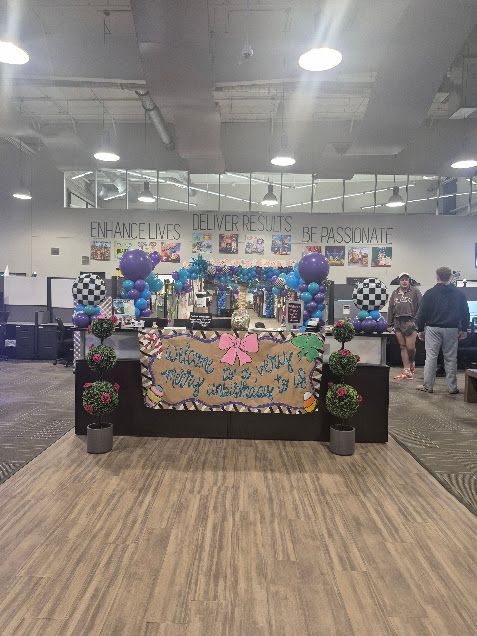 Decorated booth with colorful balloons and a banner at an indoor event hall, with people nearby