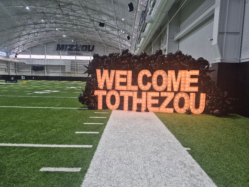 Indoor football field with a large glowing sign reading “WELCOME TO THE ZOU” beside the sideline.