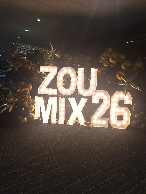 Illuminated “ZOU MIX26” sign with gold balloons in a dark event space