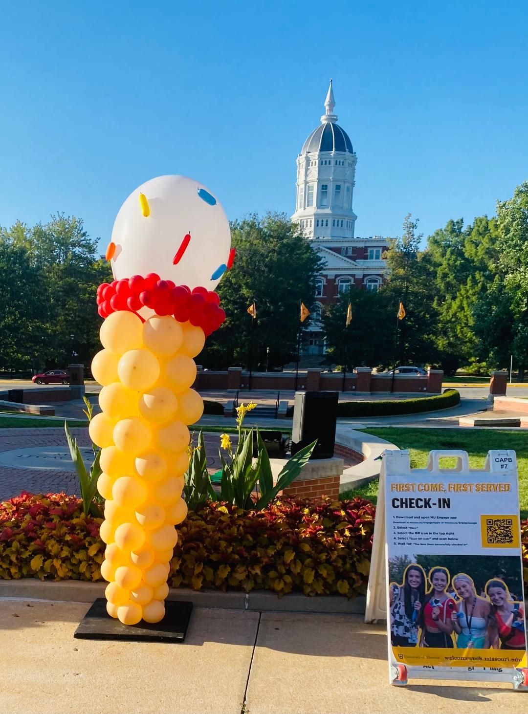 Colorful balloon display in a park with a white-domed building and event sign in the background