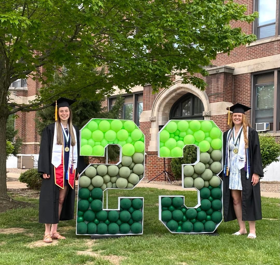 Two graduates standing beside a large green-and-silver “23” balloon display on a lawn by a brick building.