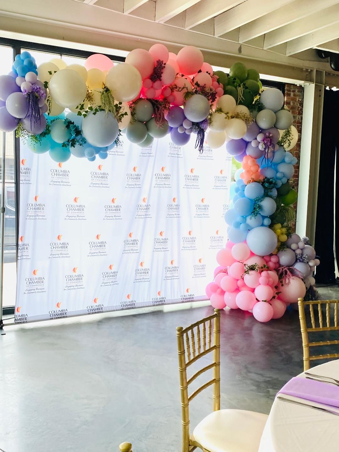 Colorful balloon arch and backdrop at a decorated event space with gold chairs and a lavender tablecloth