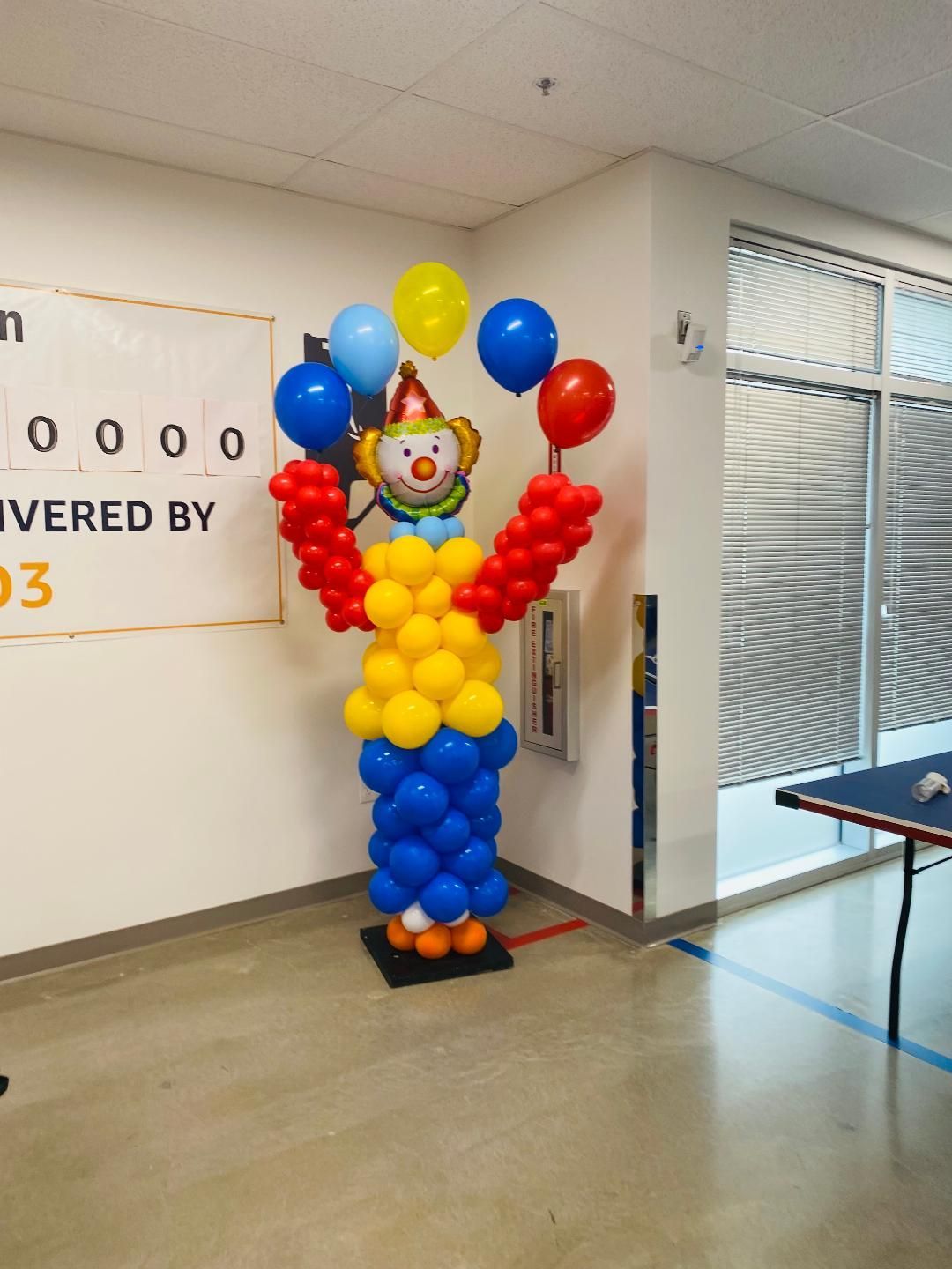 Balloon figure in red, yellow, and blue standing in a room beside a table and window.