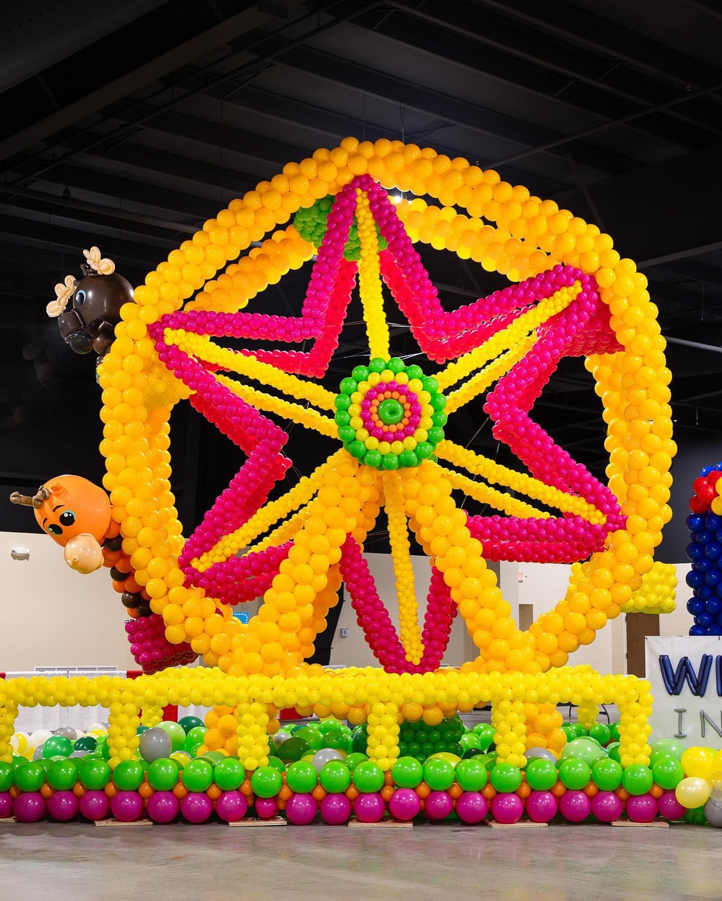Colorful balloon Ferris wheel display with a yellow frame and pink star center at an indoor event