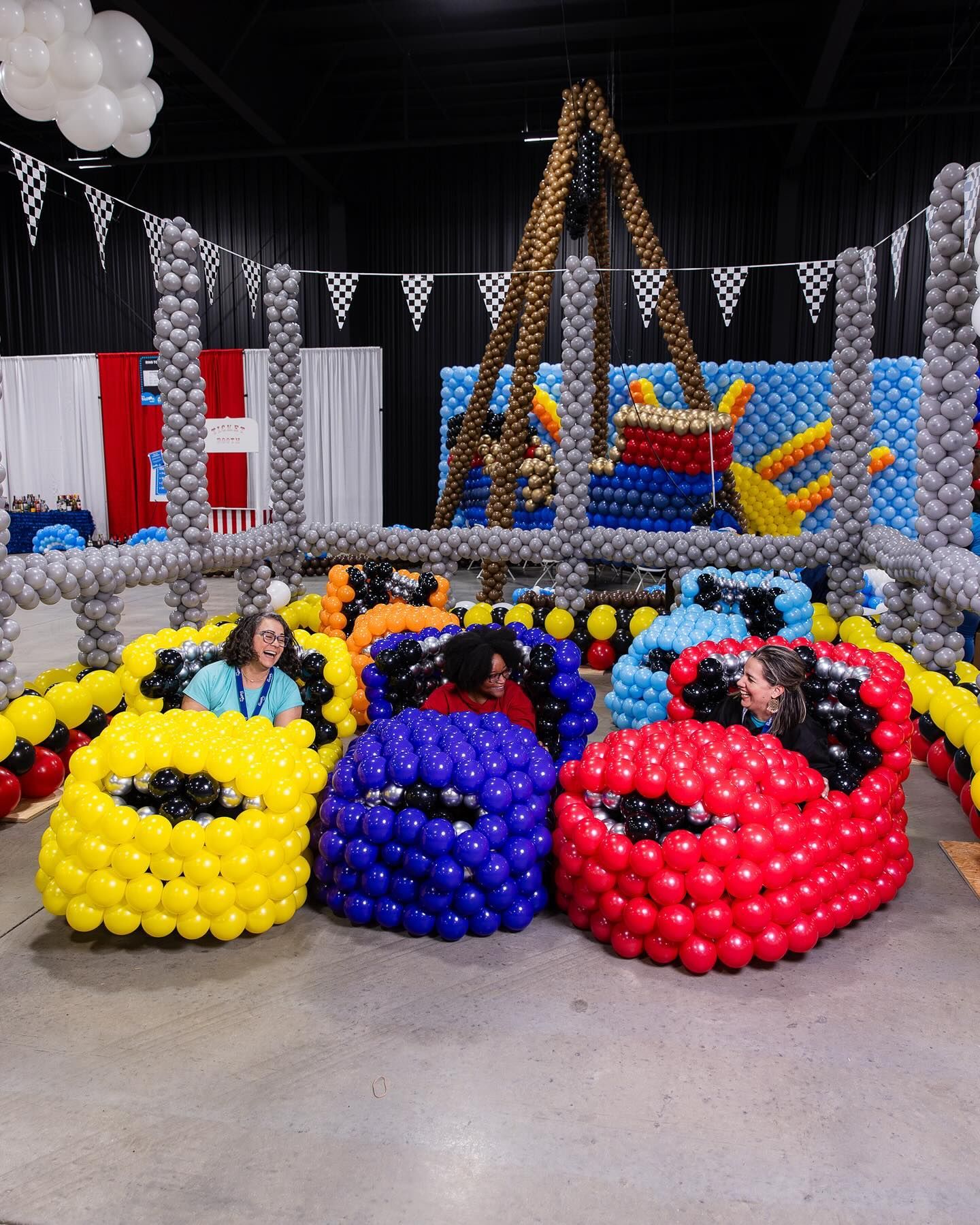 Balloon car display with three colorful cars and people seated inside at an indoor event