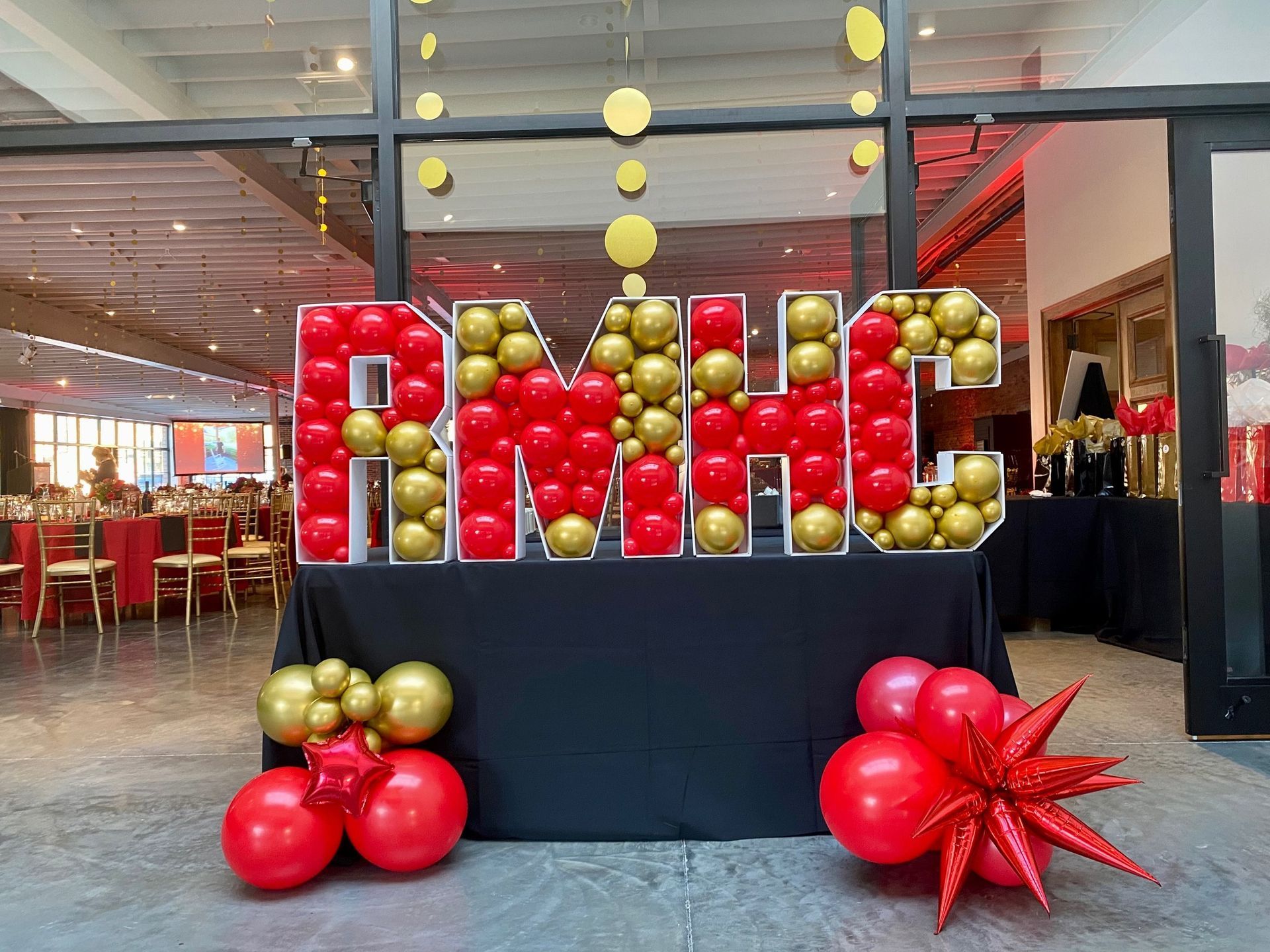 Red and gold balloon letters spelling “RMC” displayed on a black pedestal in a restaurant lobby.